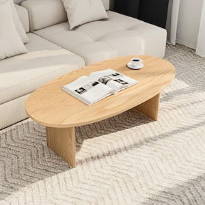 May include: A light-colored, oval wooden coffee table with a book and a cup of coffee on top. The table is supported by two vertical wooden supports. The table is placed on a patterned rug, near a beige sofa.