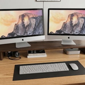 May include: Two silver iMac computers with mountain landscape screens sit on a dark wood monitor stand. A keyboard, mouse, and black mousepad are in front. A camera and a book are on the stand. The desk is made of light wood.