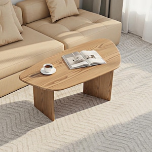 May include: A light brown wooden coffee table with a rounded top and two rectangular legs. A cup of coffee on a white saucer and an open book rest on the table. A beige sectional sofa is in the background.