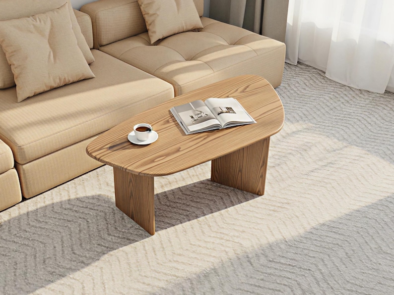 May include: A light brown wooden coffee table with a rounded top and two rectangular legs. A cup of coffee on a white saucer and an open book rest on the table. A beige sectional sofa is in the background.