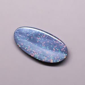 May include: An oval-shaped cabochon gemstone with a dark blue, polished surface. The stone features a scattered pattern of small, iridescent flecks in shades of pink, orange, and green that shimmer across the smooth, rounded top surface.