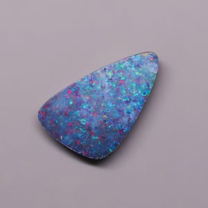 May include: A triangular-shaped loose gemstone featuring a deep blue base color with iridescent flecks of bright cyan, electric blue, and small spots of vibrant red and pink throughout the surface, creating a multi-colored speckled appearance.