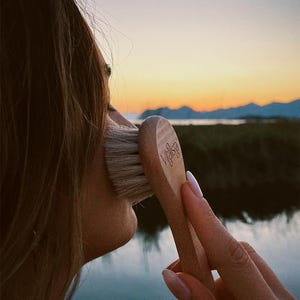 Può includere: Una spazzola per il viso in legno con la scritta "Vitalsia" stampata sul manico. La spazzola viene utilizzata sul viso. Lo sfondo presenta uno specchio d'acqua e un tramonto.