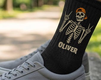 May include: A black crew sock featuring a graphic of a skeleton wearing an orange baseball cap and making a peace sign with both hands. The name OLIVER is printed in a bold, light-colored font below the skeleton graphic on the side of the sock.