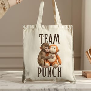 May include: A cream-colored tote bag with the words "TEAM PUNCH" printed in dark brown. The bag features an illustration of two monkeys hugging. The bag is held by a person. A necklace is displayed on a wooden bust.