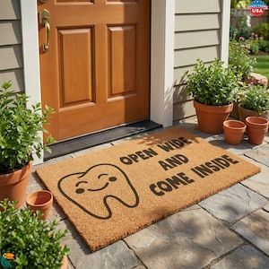May include: A rectangular doormat made of natural fibers, featuring a smiling tooth graphic and the words "OPEN WIDE AND COME INSIDE" in black. The mat is placed in front of a wooden door, with potted plants on either side.