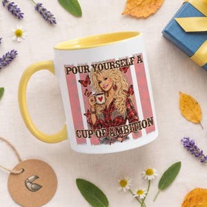 May include: A white ceramic mug with a yellow handle and interior, featuring a graphic of a blonde woman in a red plaid shirt holding a heart-adorned mug, surrounded by butterflies and the text "POUR YOURSELF A CUP OF AMBITION" in brown lettering.