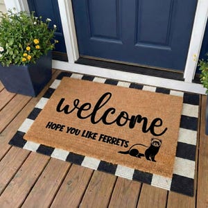 May include: A rectangular doormat with a brown center and a black and white striped border. The mat features the word "welcome" in black script, along with the phrase "HOPE YOU LIKE FERRETS" and a ferret illustration.