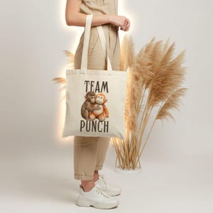 May include: Cream-colored tote bag with the words "TEAM PUNCH" in black block letters. The bag features an illustration of two hugging monkeys. The bag has long, cream-colored handles.