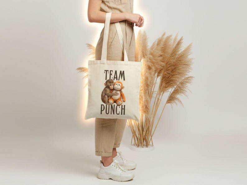 May include: Cream-colored tote bag with the words "TEAM PUNCH" in black block letters. The bag features an illustration of two hugging monkeys. The bag has long, cream-colored handles.