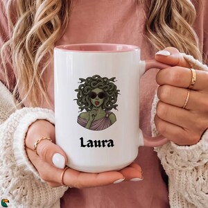 May include: A white ceramic mug with a pink interior and handle, featuring a graphic illustration of a green-skinned figure with snakes for hair, wearing sunglasses and holding a red lollipop, with the name Laura printed in black text below.