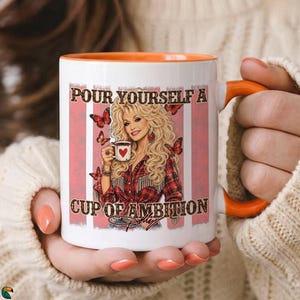 May include: A white ceramic mug with an orange interior and handle, featuring a graphic of a blonde woman in a red plaid shirt holding a coffee cup. The text reads, "POUR YOURSELF A CUP OF AMBITION" with butterflies and a signature at the bottom.