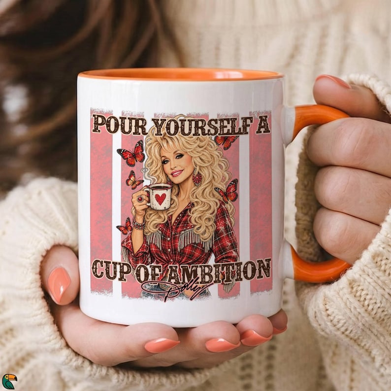 May include: A white ceramic mug with an orange interior and handle, featuring a graphic of a blonde woman in a red plaid shirt holding a coffee cup. The text reads, "POUR YOURSELF A CUP OF AMBITION" with butterflies and a signature at the bottom.