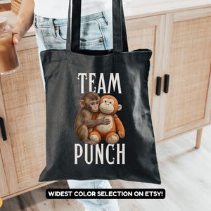May include: Black tote bag featuring the words "TEAM PUNCH" in white, with an illustration of two hugging monkeys. The bag is held by a person. The words "WIDEST COLOR SELECTION ON ETSY!" are at the bottom.