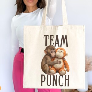 May include: Cream-colored tote bag featuring the words "TEAM PUNCH" in dark brown letters. The bag displays a graphic of two hugging monkeys, one brown and one orange. The bag is held up by a person.