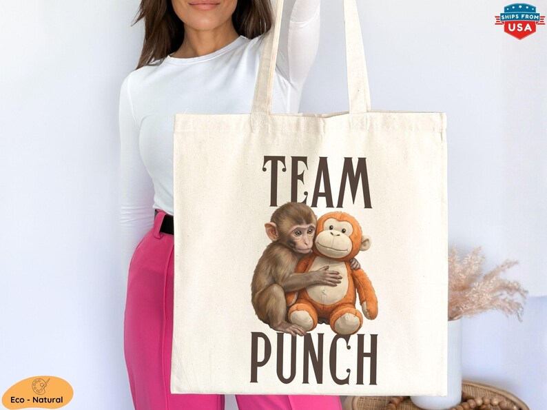 May include: Cream-colored tote bag featuring the words "TEAM PUNCH" in dark brown letters. The bag displays a graphic of two hugging monkeys, one brown and one orange. The bag is held up by a person.
