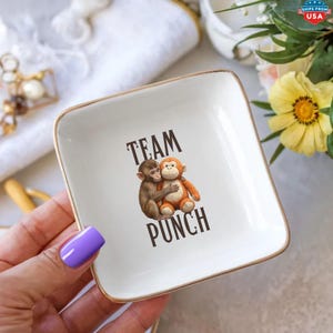 May include: A square white ceramic trinket dish with a gold-toned rim. The center features a graphic illustration of a small brown monkey hugging an orange stuffed monkey toy, with the dark brown text "TEAM PUNCH" printed above and below the figures.