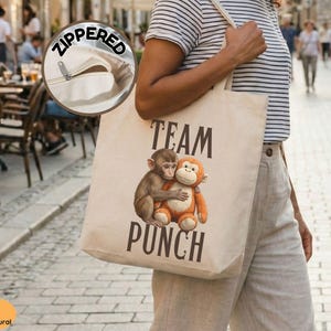 May include: A natural canvas tote bag with a zipper closure. The bag features a graphic of two hugging monkeys and the words "TEAM PUNCH" in black. The bag is carried by a person.