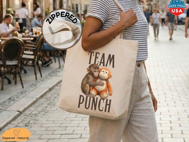 May include: A natural canvas tote bag with a zipper closure. The bag features a graphic of two hugging monkeys and the words "TEAM PUNCH" in black. The bag is carried by a person.