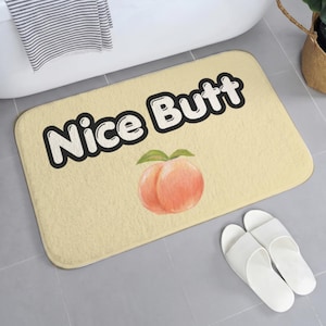 May include: A rectangular bath mat with a light beige surface featuring the text Nice Butt in bold black letters with a white outline. Below the text is a centered illustration of a peach with two green leaves, rendered in soft orange and pink tones.
