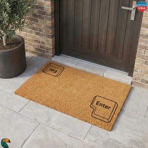May include: A rectangular tan coir doormat featuring two black graphic outlines of computer keyboard keys. The top left key is labeled with the text Esc, and the bottom right key is labeled with the text Enter, both in a bold black font.