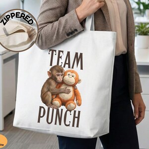 May include: White tote bag with a zippered pocket, featuring the text "TEAM PUNCH" in brown letters. The design includes two hugging monkeys, one brown and one orange. The bag is carried by a person.