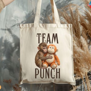 May include: Cream-colored tote bag with the words "TEAM PUNCH" in dark brown letters. The bag features a graphic of two monkeys hugging. One monkey is brown, and the other is orange. The bag has two cream-colored handles.