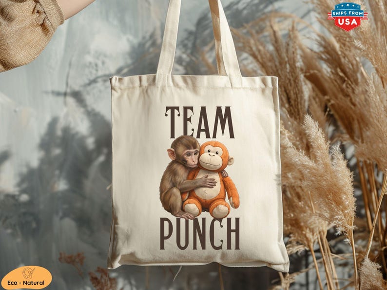 May include: Cream-colored tote bag with the words "TEAM PUNCH" in dark brown letters. The bag features a graphic of two monkeys hugging. One monkey is brown, and the other is orange. The bag has two cream-colored handles.