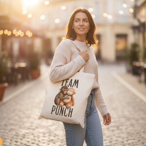 May include: A cream-colored tote bag with the words "TEAM PUNCH" printed in black. The bag features an illustration of two monkeys hugging. The person is wearing a cream sweater and blue jeans.