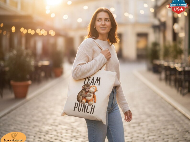 May include: A cream-colored tote bag with the words "TEAM PUNCH" printed in black. The bag features an illustration of two monkeys hugging. The person is wearing a cream sweater and blue jeans.
