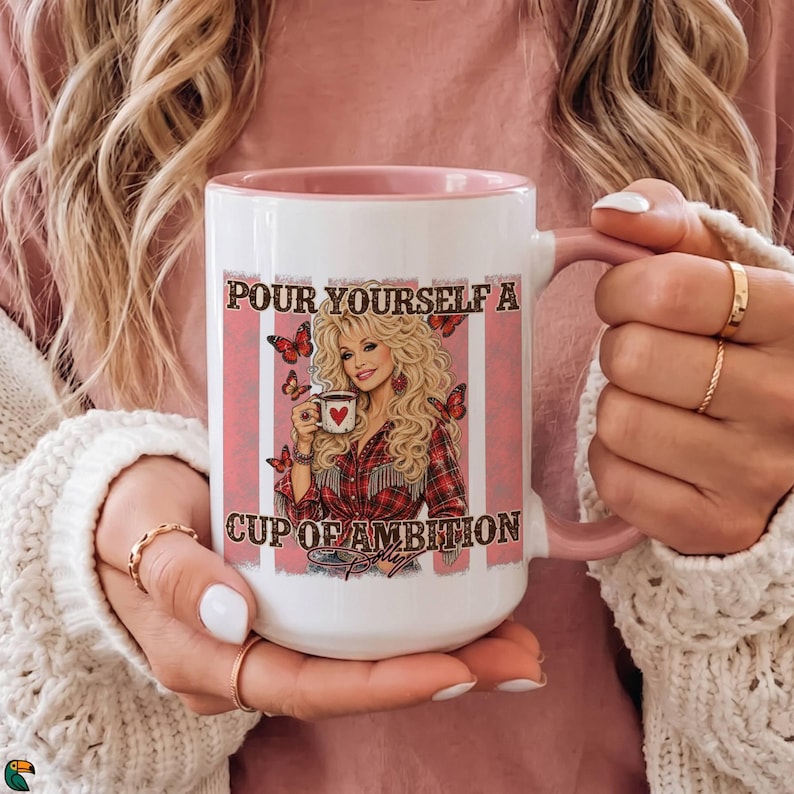 May include: A white ceramic mug with a light pink interior and handle. The front features a graphic of a blonde woman in a red plaid shirt holding a small mug with a heart, surrounded by butterflies and the text "POUR YOURSELF A CUP OF AMBITION".