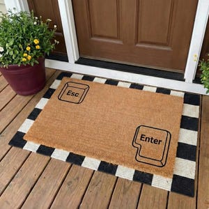 May include: A rectangular doormat made of natural fibers, featuring the words "Esc" and "Enter" in black, resembling computer keys. The mat is bordered by a black and white striped pattern and sits on a wooden porch.