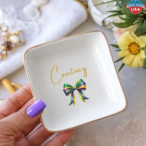 May include: A square, white ceramic trinket dish with a gold rim. The dish features the name "Courtney" in gold script and a colorful bow design in purple, green, and yellow. The dish is held by a person with purple nail polish.