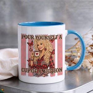 May include: A white ceramic mug with a bright blue interior and handle. The front features a graphic of a blonde woman in a red plaid shirt holding a coffee cup with a heart, surrounded by butterflies and the text Pour Yourself A Cup Of Ambition.