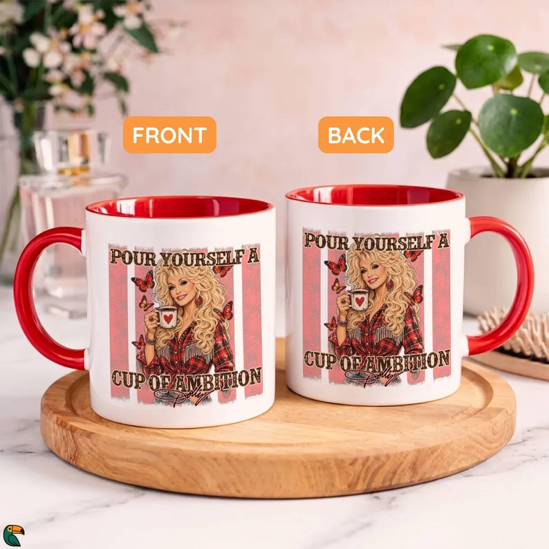 May include: Two white ceramic mugs with red interiors and handles. The mugs feature a graphic of a blonde woman in a plaid shirt holding a mug, surrounded by butterflies and the text "POUR YOURSELF A CUP OF AMBITION" on a pink striped background.