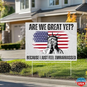 May include: A white yard sign with the text "ARE WE GREAT YET? BECAUSE I JUST FEEL EMBARRASSED." The sign features the American flag and an illustration of the Statue of Liberty covering her face. The sign is displayed on a green lawn.