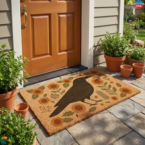 May include: A rectangular doormat with a crow design. The mat is tan with a pattern of sunflowers and green leaves. The crow is dark brown and is facing left. The doormat is placed in front of a brown wooden door.