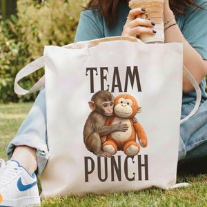 May include: A white canvas tote bag with the words "TEAM PUNCH" in dark brown letters. The bag features an illustration of two hugging monkeys, one brown and one orange. The bag has white handles.