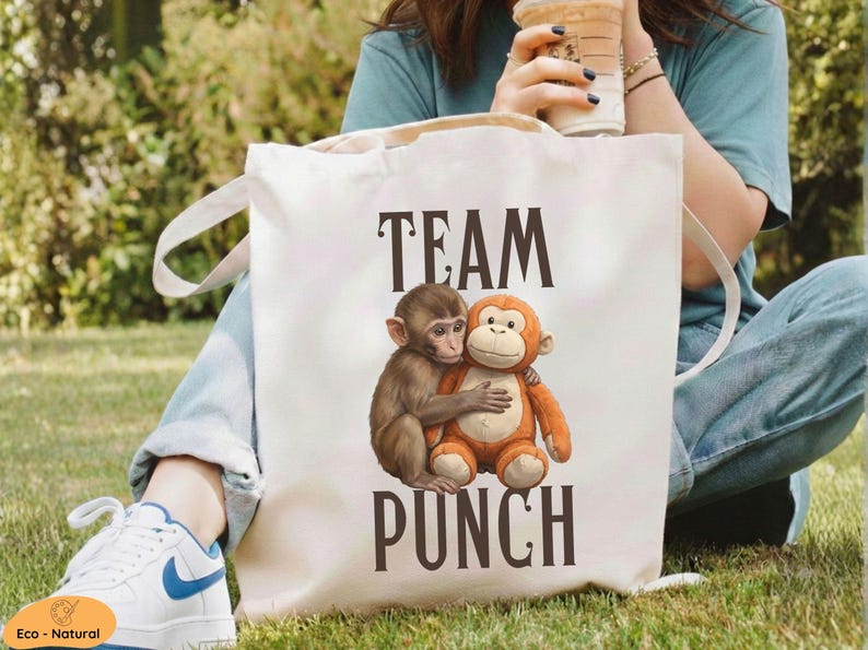 May include: A white canvas tote bag with the words "TEAM PUNCH" in dark brown letters. The bag features an illustration of two hugging monkeys, one brown and one orange. The bag has white handles.