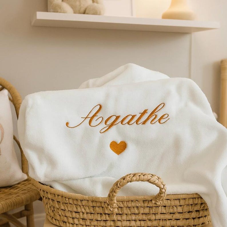 May include: A white, plush baby blanket with the name "Agathe" embroidered in gold script, along with a small gold heart. The blanket is folded and placed in a woven basket, suggesting a nursery or baby-themed product.