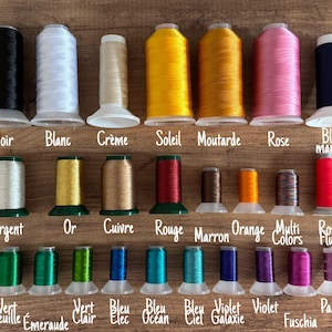 May include: A collection of colorful thread spools in various shades, including black, white, yellow, pink, and blue. Each spool is labeled with its color name, such as "Noir" and "Rose". The spools are arranged on a wooden surface.