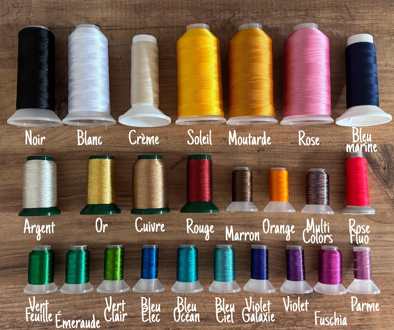 May include: A collection of colorful thread spools in various shades, including black, white, yellow, pink, and blue. Each spool is labeled with its color name, such as "Noir" and "Rose". The spools are arranged on a wooden surface.