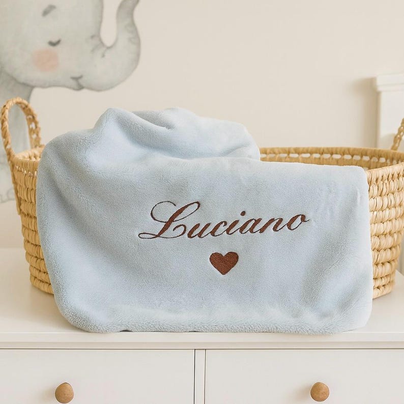 May include: A light blue baby blanket embroidered with the name "Luciano" in brown script, along with a small brown heart. The blanket is folded over a woven basket, suggesting a nursery setting.