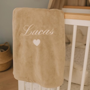 May include: A tan baby blanket with the name "Lucas" embroidered in white script, along with a white heart. The blanket is draped over a white crib, suggesting a nursery setting. The soft, plush texture is visible.