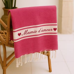 Peut inclure: Une serviette fuchsia avec une bande blanche portant l'inscription "Mamie d'amour" et des symboles de cœur. La serviette a une frange le long du bord inférieur et est drapée sur une chaise en osier.