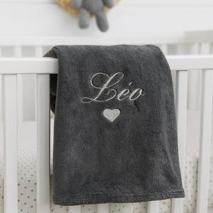 May include: A dark gray baby blanket with the name "L&eacute;o" embroidered in white script, along with a silver heart. The blanket is draped over a white crib rail. A stuffed animal is visible in the background.