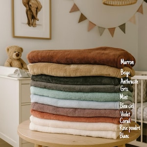 May include: A stack of ten plush, folded towels in various colors, including brown, beige, gray, and blue. The towels are neatly arranged on a wooden table, with color names labeled on the side. The background includes a framed elephant print and a teddy bear.