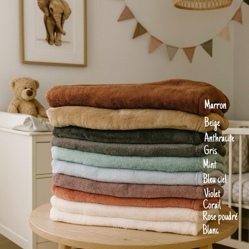 May include: A stack of ten plush, folded towels in various colors, including brown, beige, gray, and blue. The towels are neatly arranged on a wooden table, with color names labeled on the side. The background includes a framed elephant print and a teddy bear.