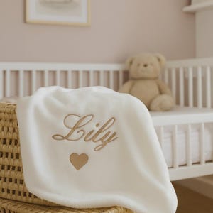 Peut inclure: Une couverture de bébé blanche et douce avec le nom "Lily" brodé en lettres dorées, accompagné d'un cœur doré. La couverture est posée sur un panier en osier, avec un lit bébé et un ours en peluche en arrière-plan.