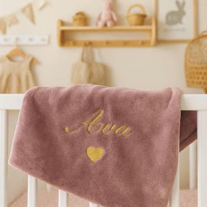 May include: A dusty rose baby blanket with the name "Ava" embroidered in gold script, along with a gold heart. The blanket is draped over a white crib rail. The nursery setting includes a wooden shelf with decor.